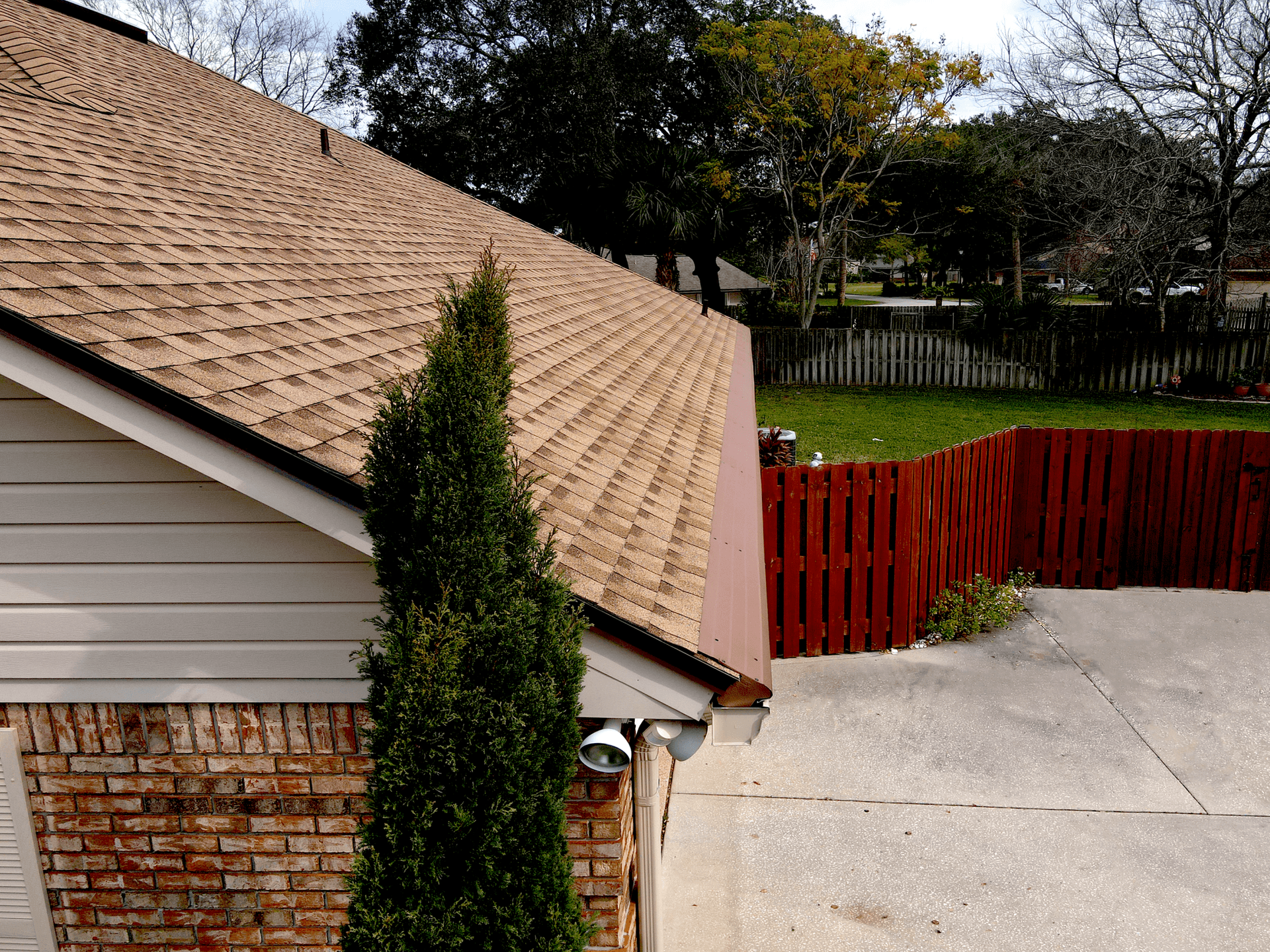 A sloped roof with brown shingles covers a brick house. A tall bush stands by the house. A wooden fence borders a green yard. Calm suburban scene.
